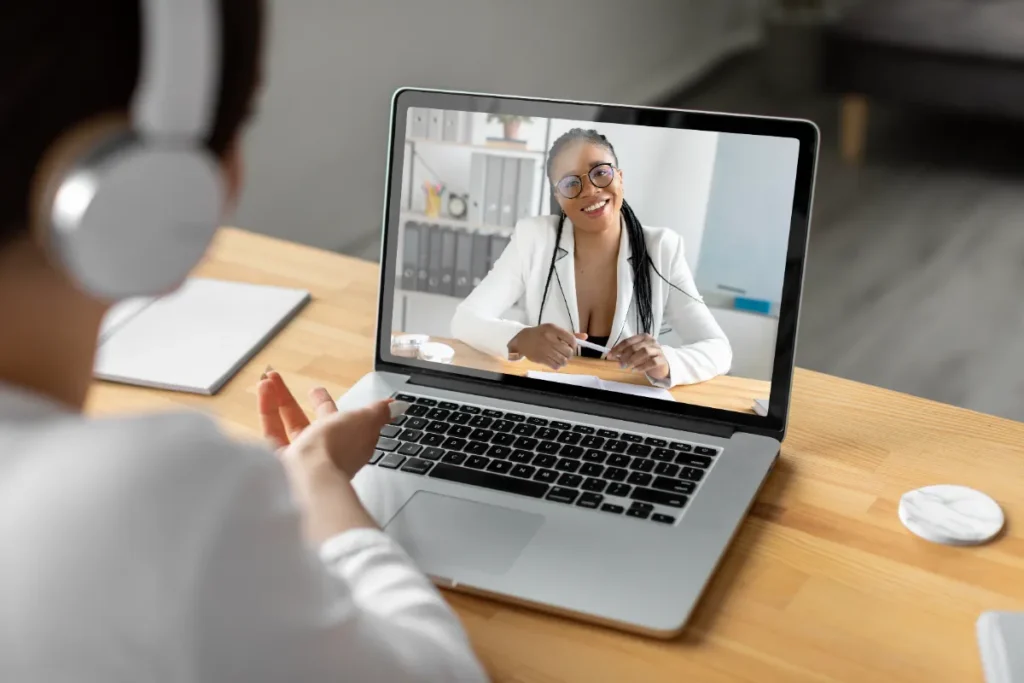 Pessoa utilizando um fone de ouvido em frente a um notebook, realizando uma teleconsulta por vídeo com uma médica sorridente, ilustrando o atendimento médico à distância.