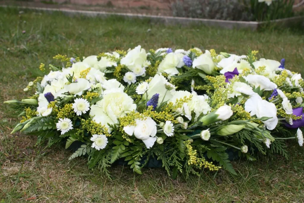 Close-up de uma coroa de flores fúnebres branca e verde, disposta sobre a grama, com flores como crisântemos brancos, cravos e folhagens, simbolizando uma homenagem em um funeral.