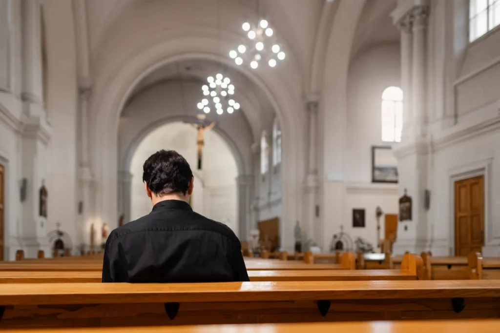 Pessoa de costas, sentada sozinha em um banco de madeira em uma igreja de arquitetura clássica, simbolizando a participação na missa de sétimo dia, um momento de acolhimento, fé e homenagem após o falecimento.
