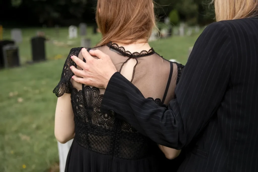Mulher de terno preto, vista por trás, abraçando outra pessoa em um cemitério. O gesto de apoio mostra o que significa "meus pêsames".