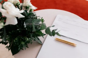 Arranjo de flores brancas ao lado de documento com canetas sobre mesa.